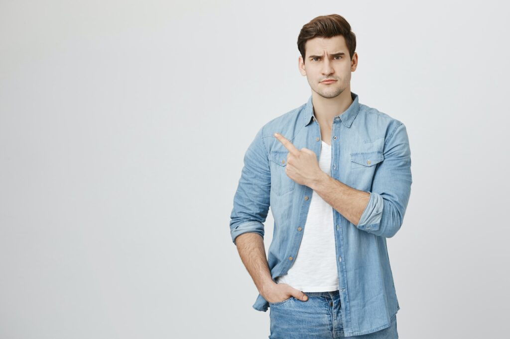 Serious looking handsome student in denim clothes squinting at camera and pointing left with index